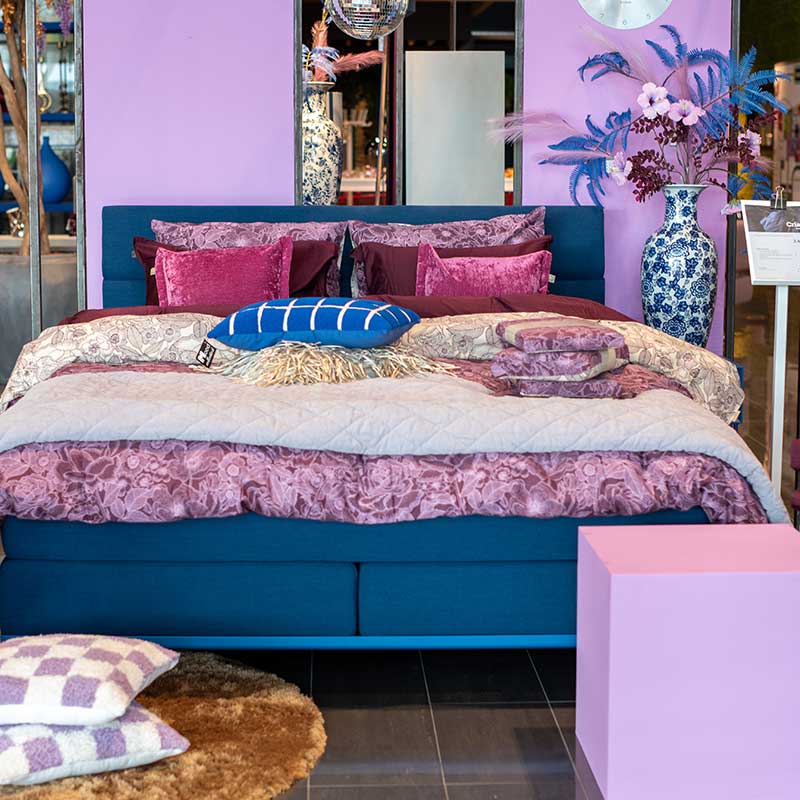 Slaapkamerdisplay met een blauw bed, paarse en roze dekens, kleurrijke kussens en decoratieve accessoires, geïnspireerd door de Cosmic Future woontrend 2025