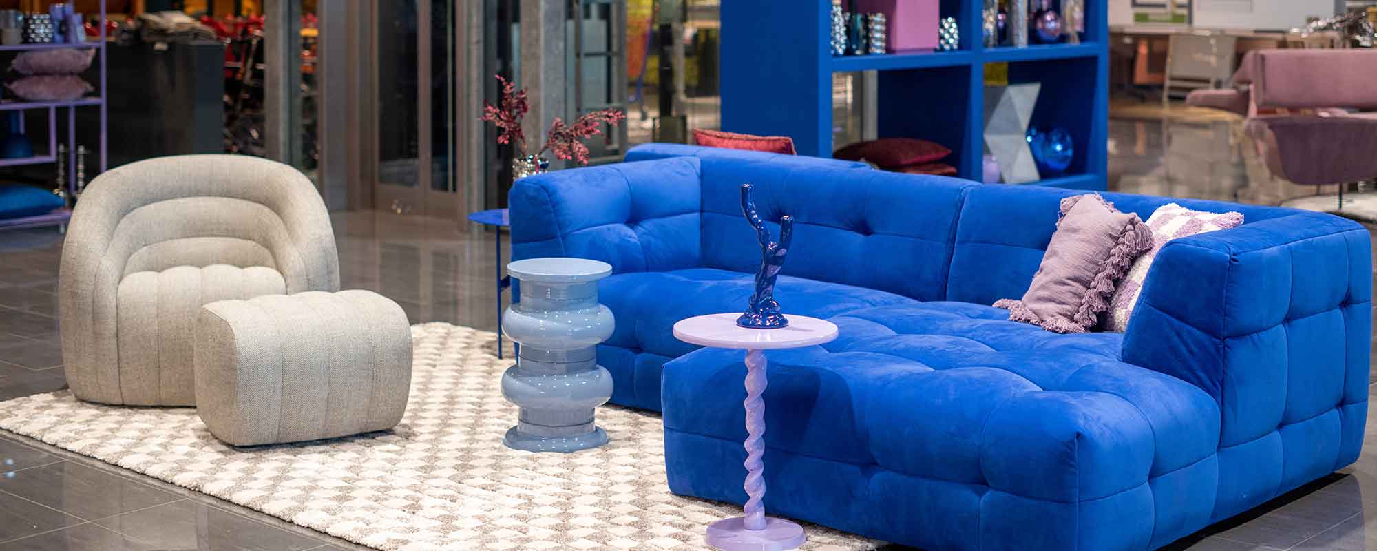 Interieursetting met een opvallende blauwe bank, moderne grijze fauteuil en kleurrijke bijzettafels, geïnspireerd door de Cosmic Future woontrend 2025.