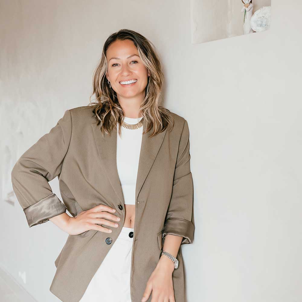 Eva van de ven, interieurstyliste, in een beige blazer, witte top, glimlachend terwijl ze tegen een muur leunt