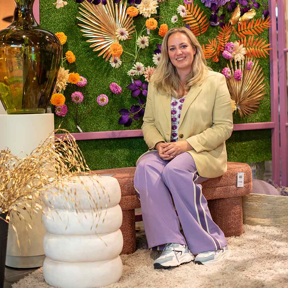 Interieur instagrammer, Torentje van Tessa, in lichtgeel jasje en paarse broek, zittend op een bankje met kleurrijke bloemenachtergrond.